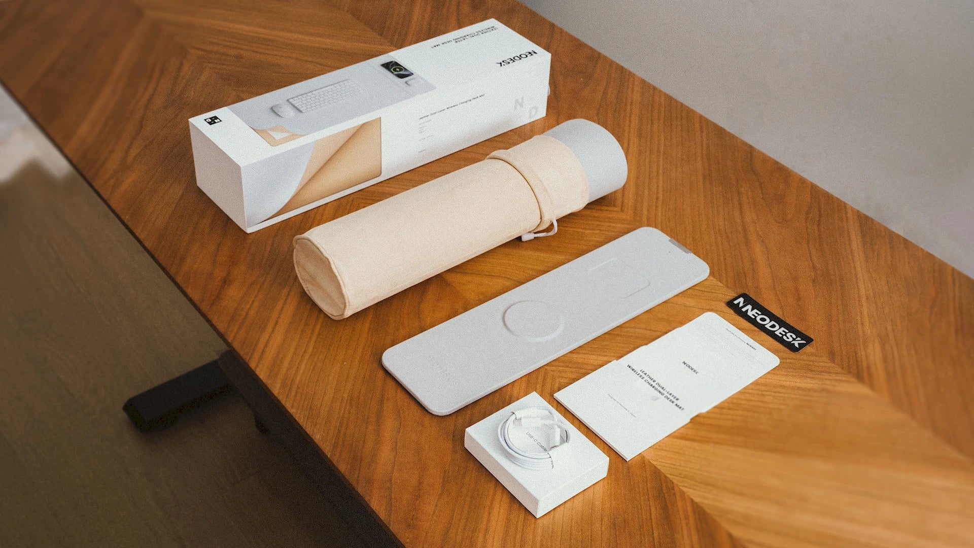 NeoDesk dual-layer charging mat package contents displayed on a wooden table, including the wireless charging mat, velvet storage bag, USB-C cable, instruction manual, and packaging box.