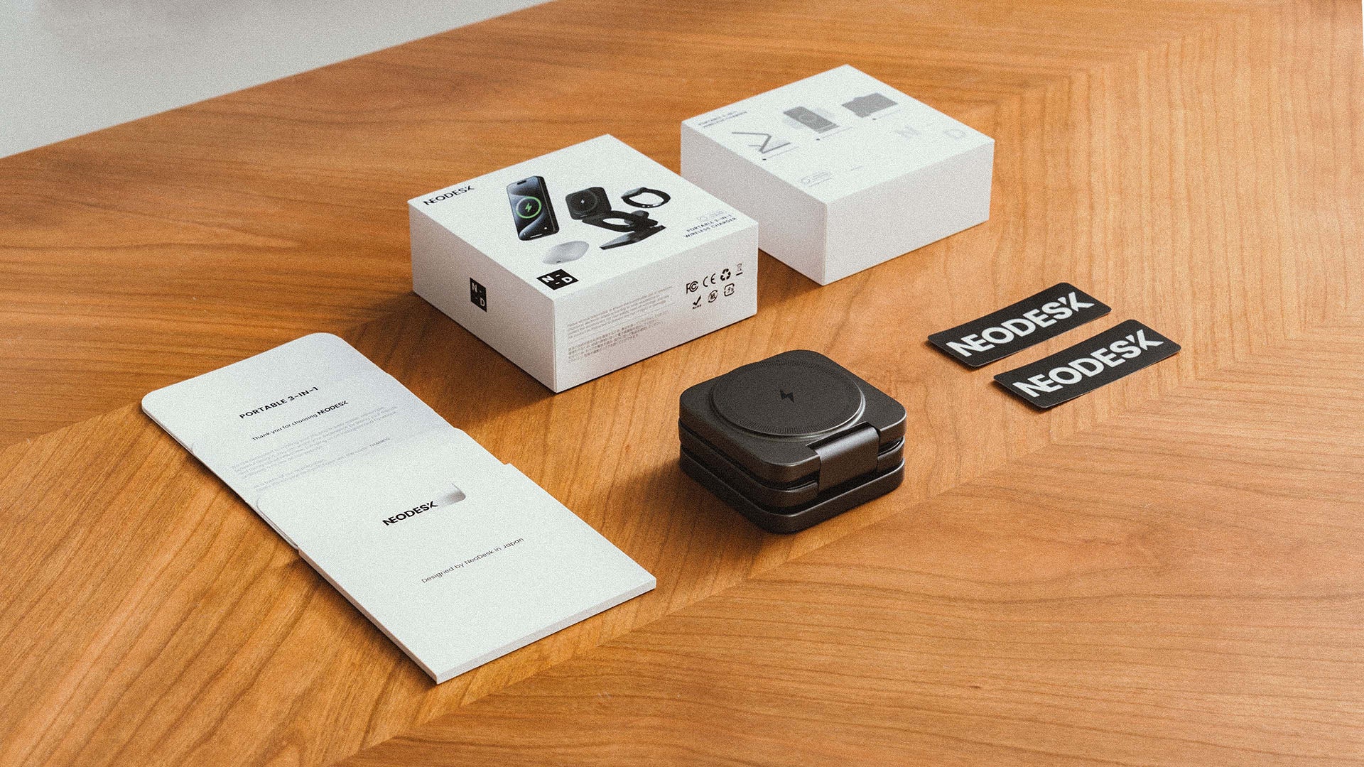 NeoDesk package contents of Portable 3-in-1 Wireless Charger with product box, folded charger, manuals, and stickers on a wooden surface